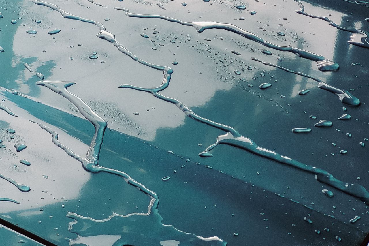 Gotas y regueros de agua sobre la pintura oscura de una carrocería, que refleja el cielo nublado.