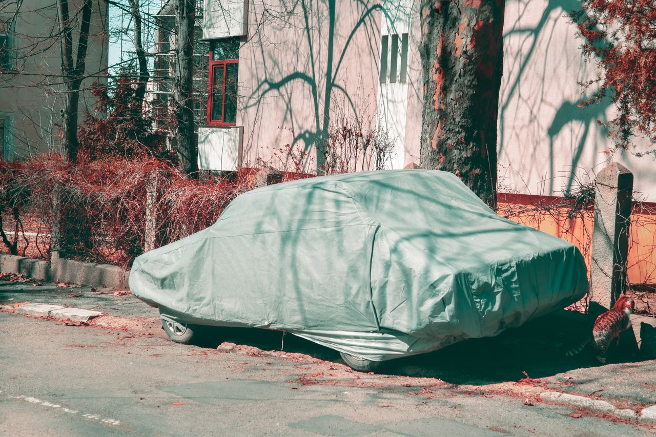 Coche aparcado junto a la acera, tapado por completo con una funda de tela clara, delante de un edificio y unos árboles.