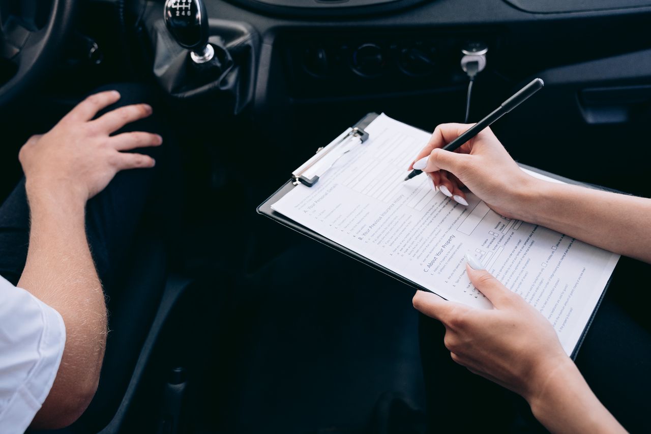 Dentro de un coche, unas manos rellenan con bolígrafo un formulario en una carpeta con pinza; al lado, el brazo de otra persona.