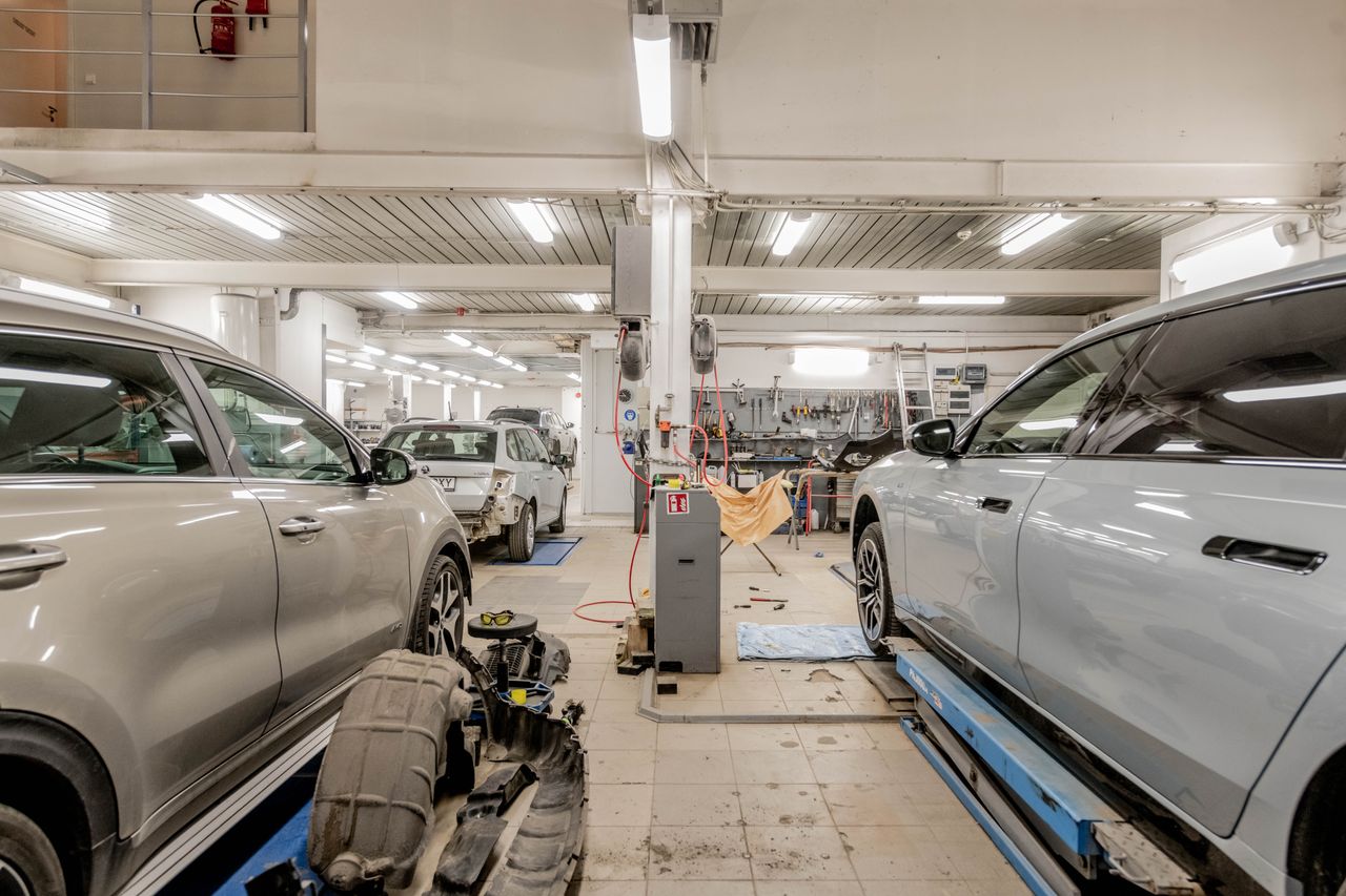 Interior de un taller con coches en reparación, uno sobre un elevador, herramientas en la pared del fondo y piezas desmontadas en el suelo.