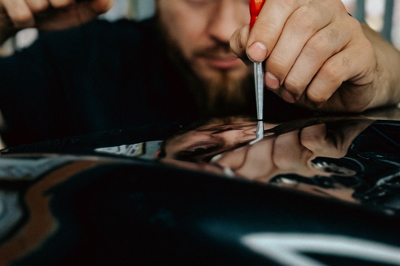 Un técnico con barba apoya la punta de una herramienta de mango rojo sobre una chapa negra y brillante en la que se refleja su cara.